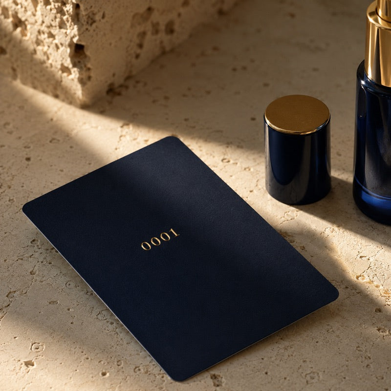 A deep navy membership card embossed with the gold number 0001, beside the navy serum bottle on cream travertine