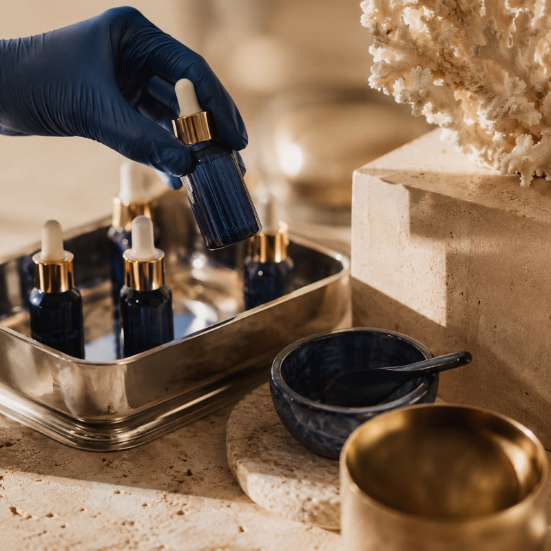 A gloved hand lifting a small navy dropper bottle from a stainless steel laboratory tray in warm light