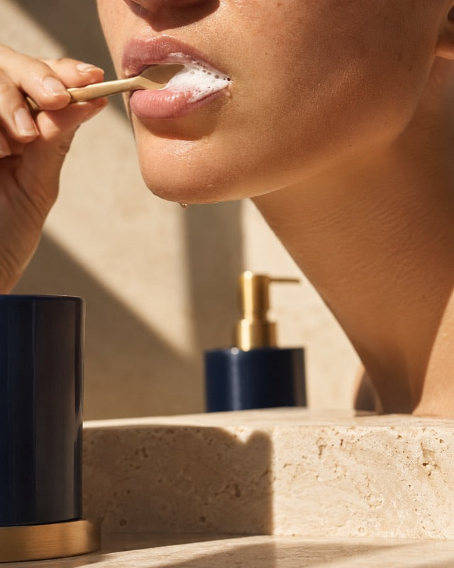 Brushing teeth with white foam gathering at the corner of the mouth, navy and gold bathroom accents in warm sunlight